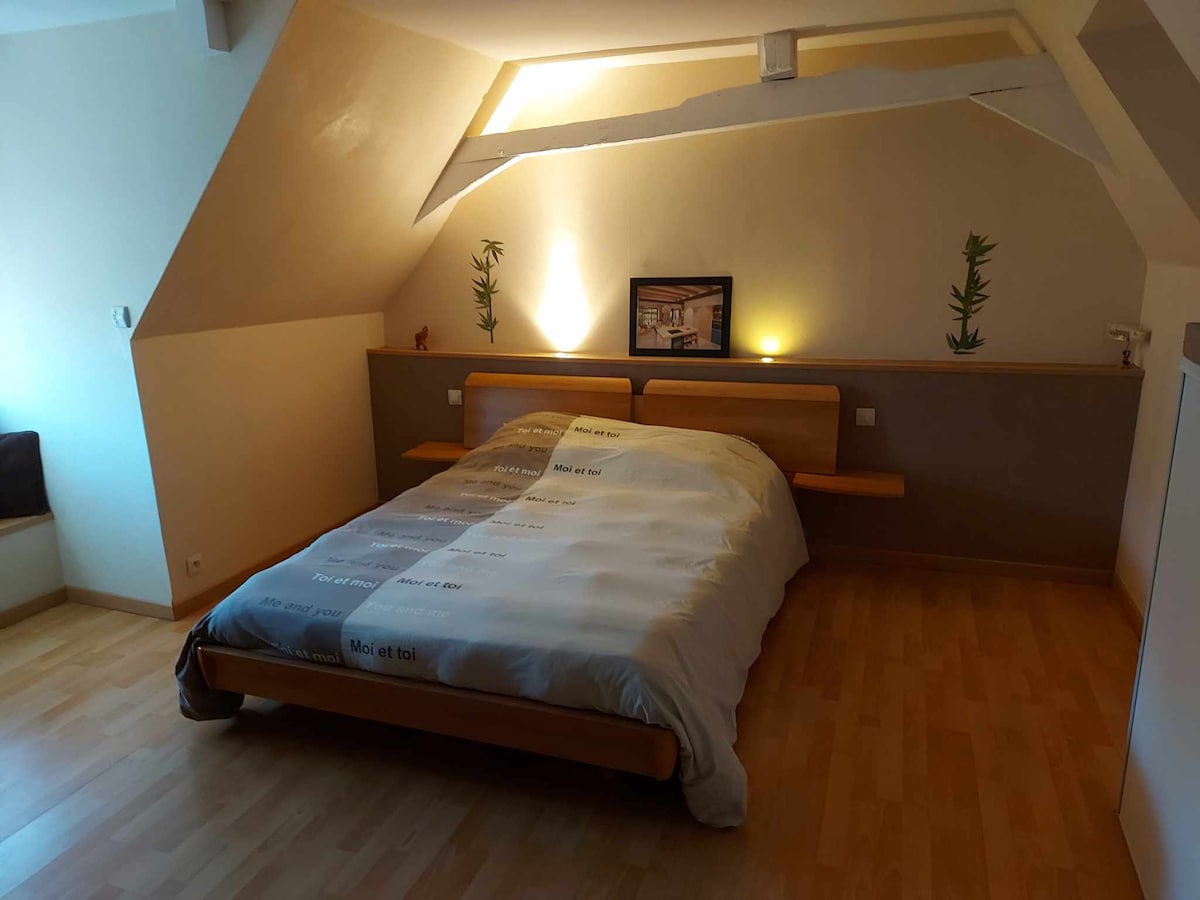A spacious bedroom features a large wooden bed with a neutral-tone comforter. Soft lighting from wall sconces creates a calm ambiance, while simple decorative elements are arranged on the walls. The wooden flooring adds warmth to the room, which has sloped ceilings enhancing the cozy feel.