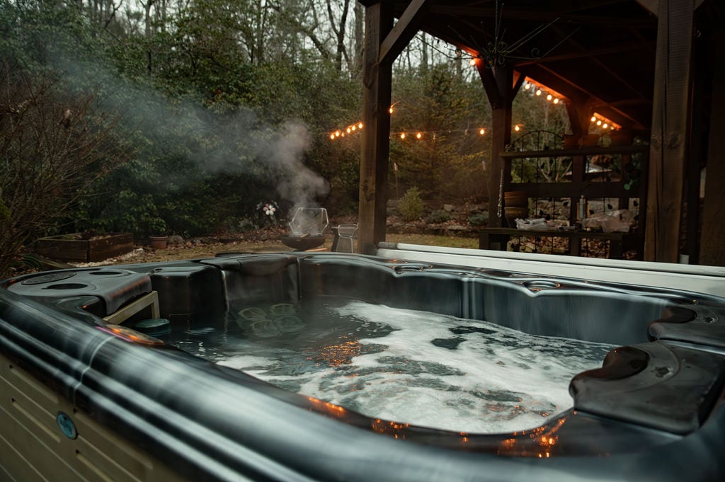 A hot tub with bubbling water is displayed in a serene outdoor setting. Steam rises gracefully above the water, and decorative string lights illuminate the surrounding area. A rustic wooden structure features shelves holding various items, creating an inviting and relaxed atmosphere.