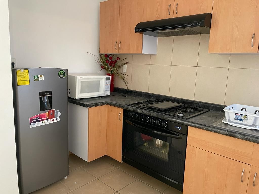 A modern kitchen features light wooden cabinets and a black marble countertop. Stainless steel appliances, including a refrigerator and a gas stove, are present. A microwave is positioned on the countertop, alongside decorative elements like a small vase with flowers.