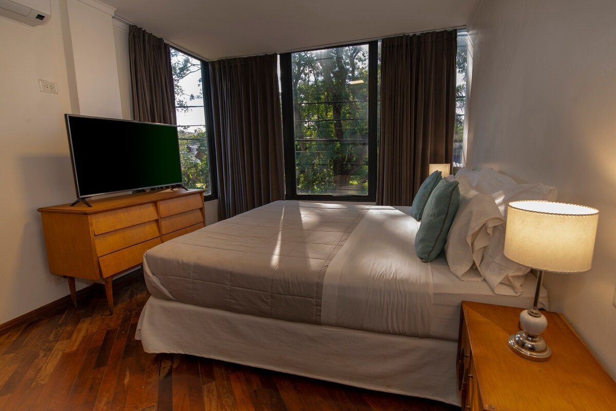 A spacious sleeping area features a large bed dressed in soft linens, accompanied by decorative pillows. A sleek wooden dresser and two bedside tables with lamps are present. Large windows allow natural light to illuminate the room, framed by flowing curtains.