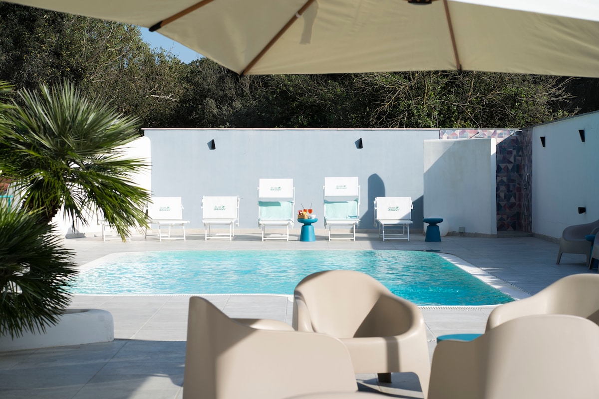 A serene outdoor space is seen featuring a refreshing swimming pool surrounded by six white lounge chairs. Nearby, two contemporary armchairs offer a comfortable seating area under the shade of a large umbrella, with greenery enhancing the tranquil environment.