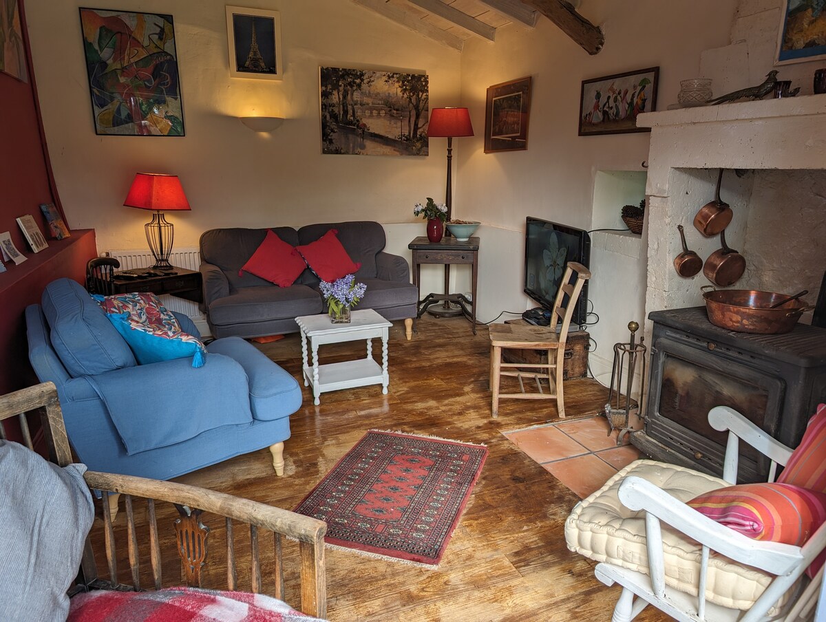 A cozy living area features a mix of seating options, including a blue armchair and a gray couch adorned with red cushions. A small white table stands centrally, and a vintage-style armchair is positioned near the fireplace. Artworks and warm lighting create a welcoming ambiance.