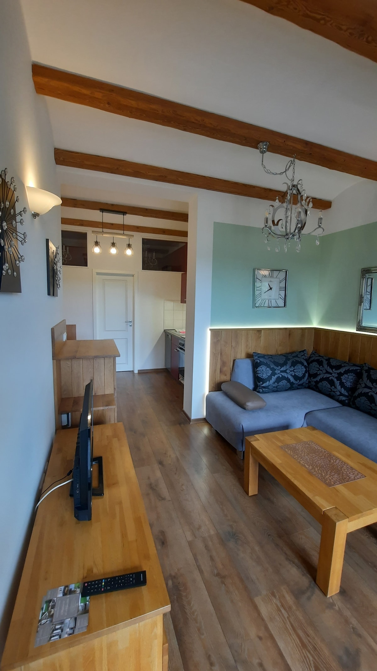 A living area features a comfortable gray sofa adorned with decorative pillows. A wooden coffee table sits in front, while a TV is positioned on a matching wooden console. The ceiling showcases exposed beams, and subtle lighting enhances a calm ambiance.