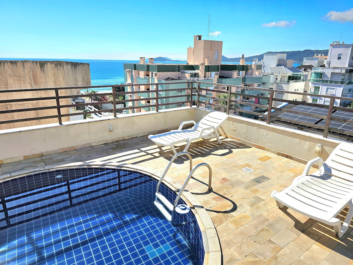 A terrace features a small blue-tiled pool, surrounded by light-colored stone tiles. Two lounge chairs are positioned for sunbathing, with an unobstructed view of the ocean and distant buildings in the background under a clear blue sky.