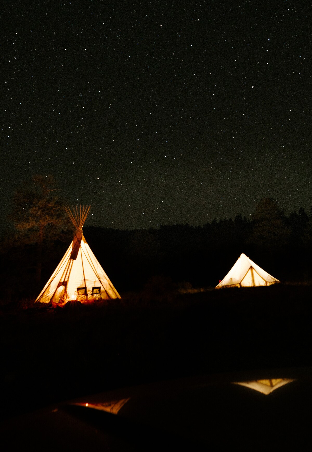 Two illuminated tipis stand under a starry night sky, their warm glow contrasting with the dark surroundings. The silhouette of the tipis features natural materials and a traditional shape, inviting a sense of adventure in the wilderness.