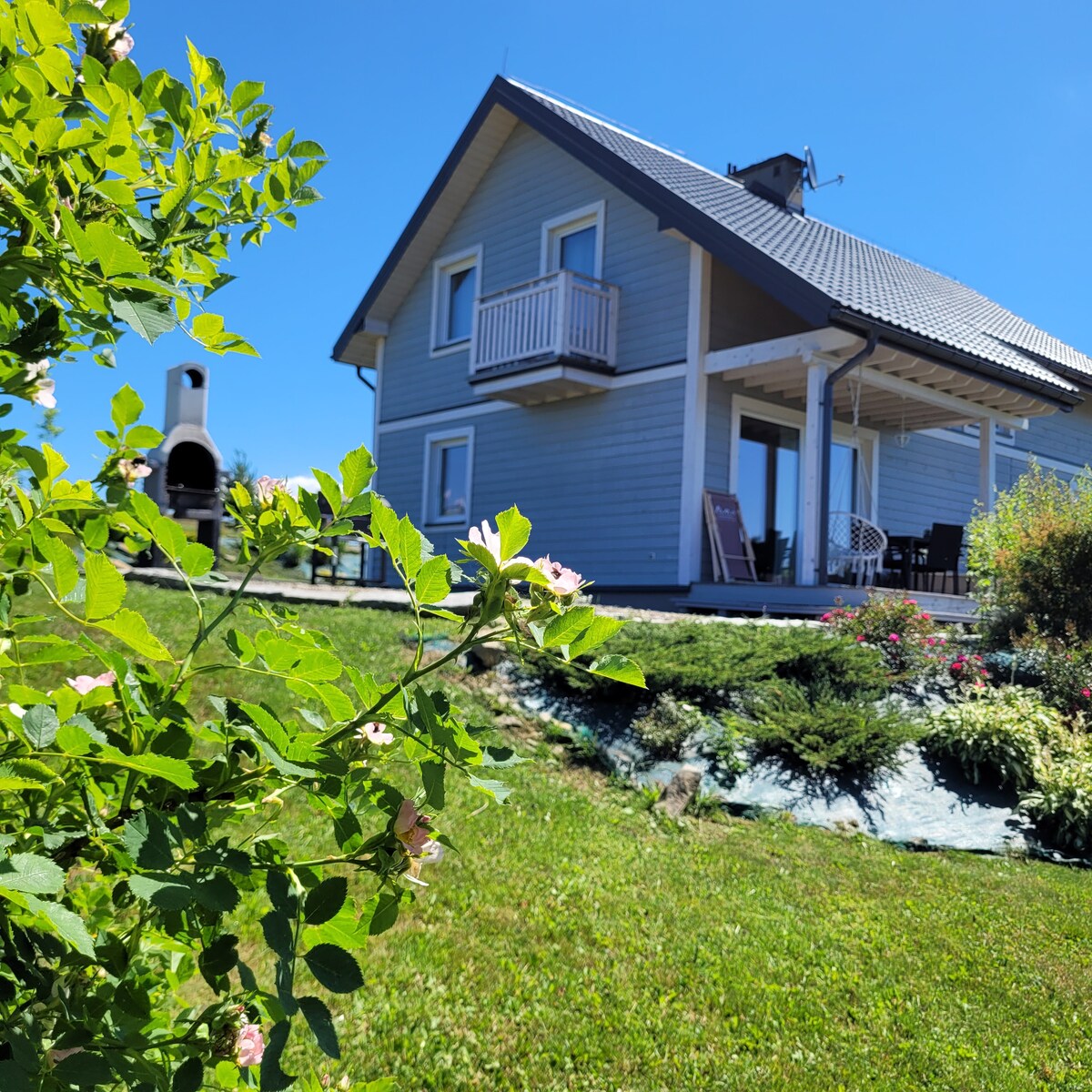 The charming wooden house is framed by vibrant greenery and flowers. A large patio extends from the house, featuring a grill and outdoor seating. The bright blue sky enhances the peaceful setting, inviting relaxation in the surrounding garden space.
