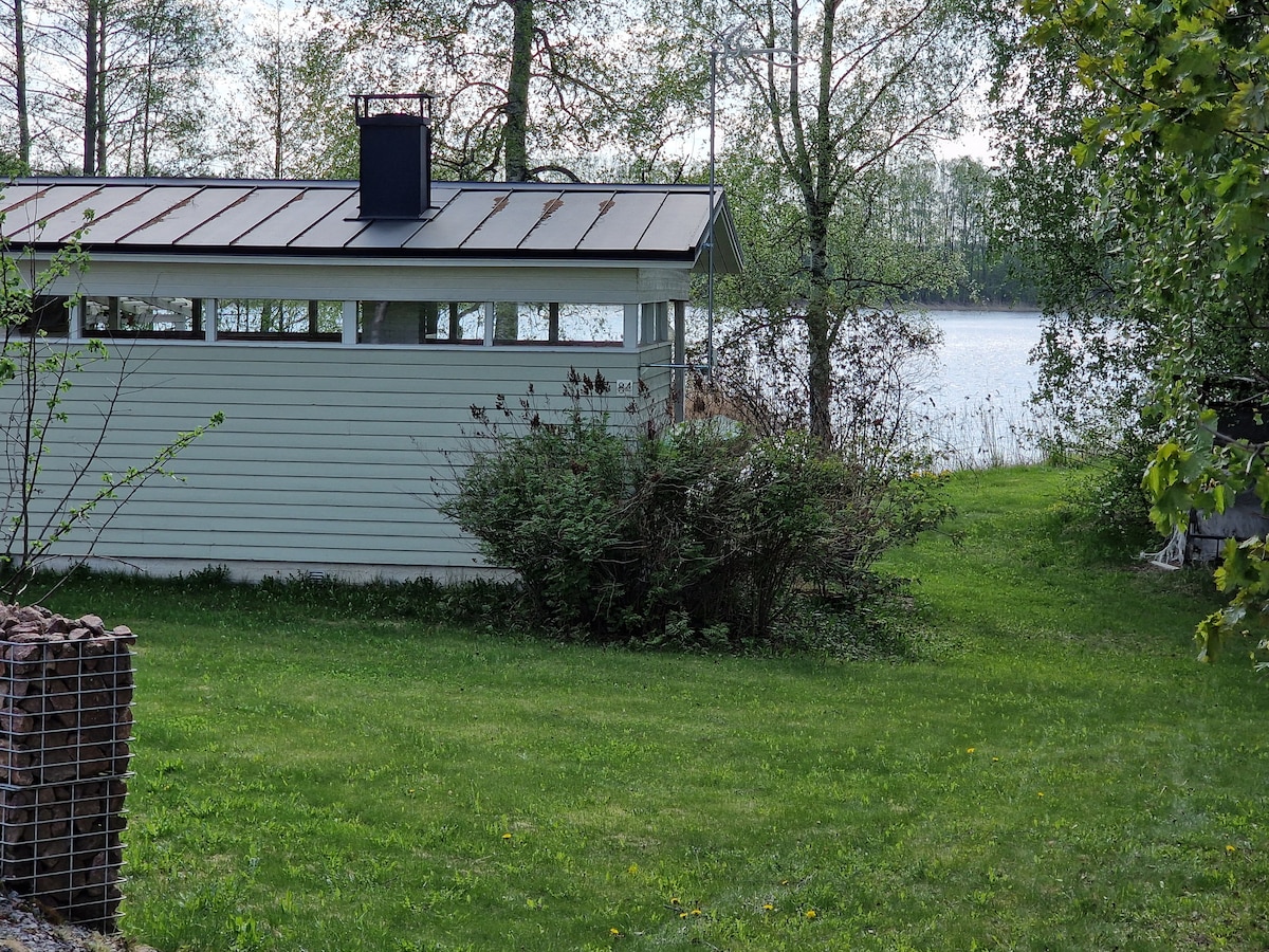 A light-colored building is situated in a serene outdoor space, surrounded by lush grass and shrubbery. The structure features expansive windows, providing views of the nearby lake, which reflects the greenery around it. The tranquil setting invites relaxation and connection with nature.