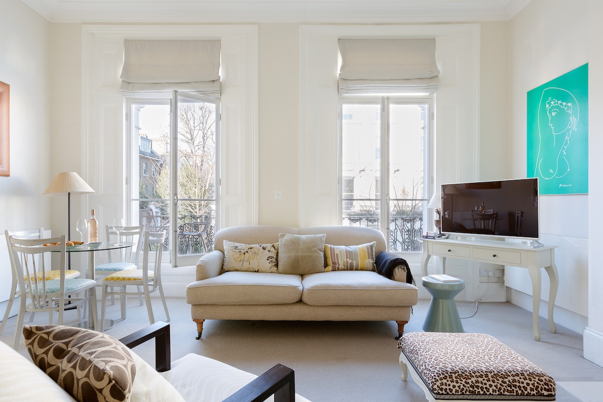 A bright living area features large windows providing ample natural light, complemented by elegant shades. A comfortable sofa and armchair surround a light coffee table, with a sleek TV on a console nearby. Artwork adds a touch of colour to the wall.
