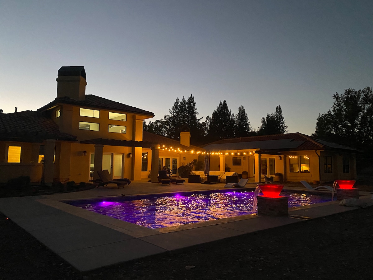 The exterior of the guest house is illuminated at dusk, showcasing a swimming pool with colorful lighting reflecting off the water. Warm lights drape over the patio area, enhancing the inviting scene surrounded by trees.