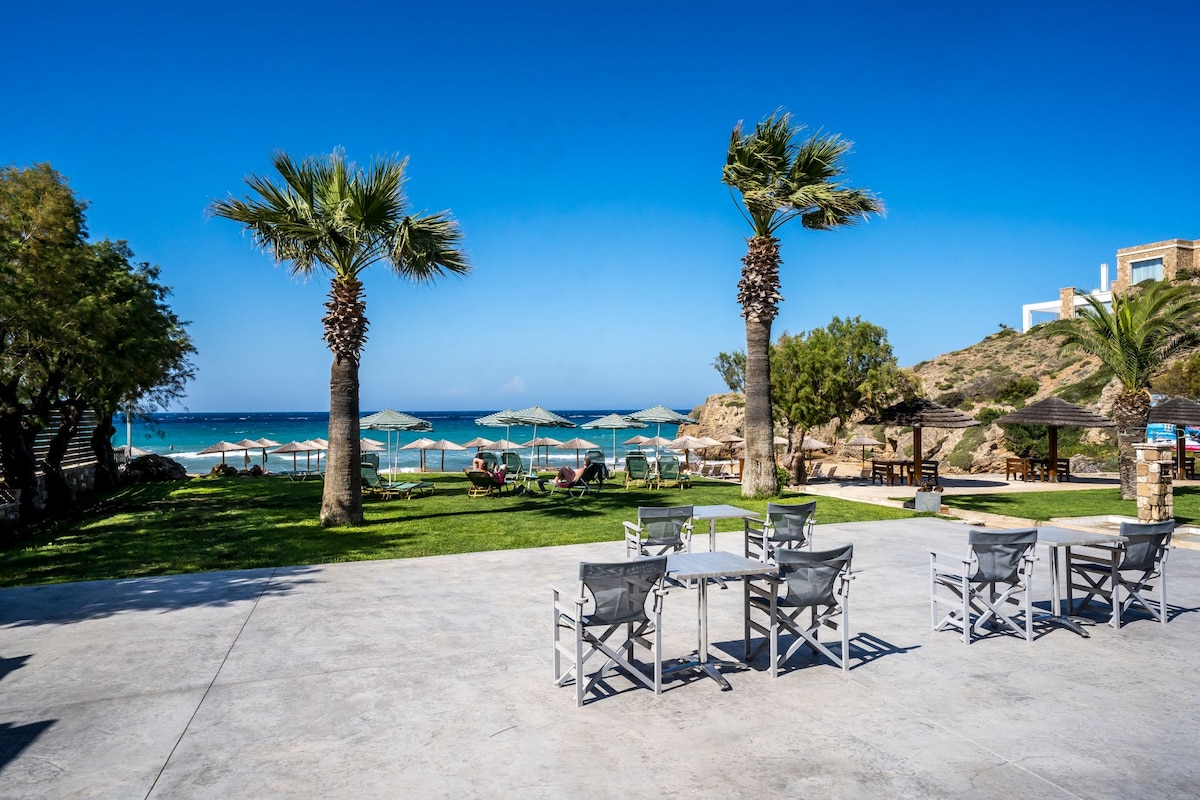 A sunlit outdoor area features neat rows of white patio tables and chairs set on a concrete surface. Lush greenery and palm trees provide shade, while sun loungers and umbrellas are arranged near the beach, where clear blue waters meet a sandy shore.