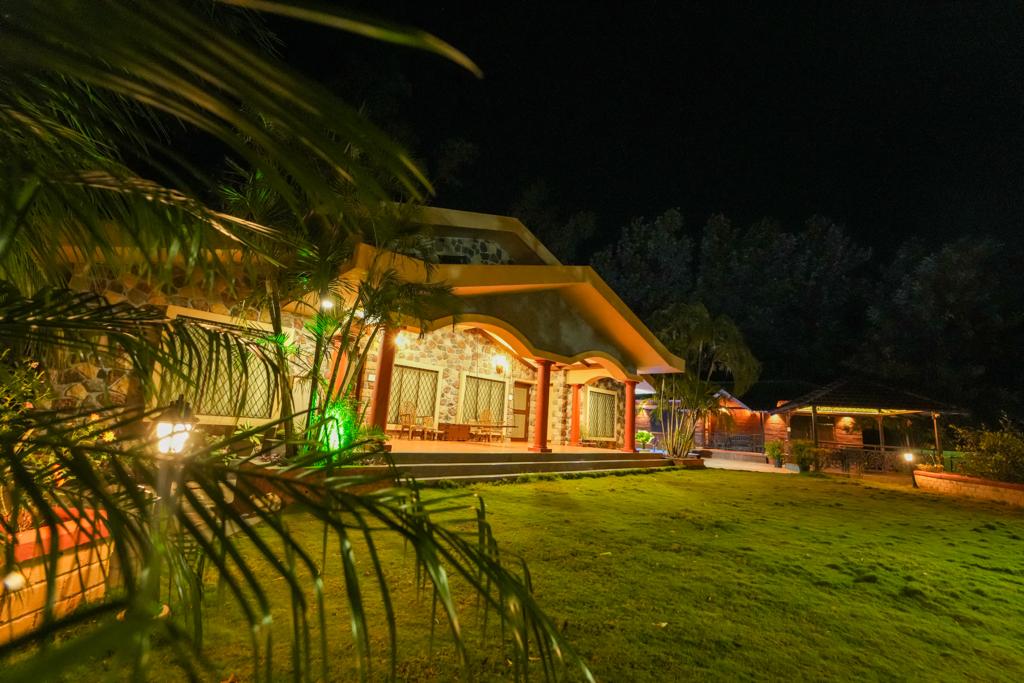 A charming villa is illuminated at night, showcasing a solid stone exterior and large arched windows. Lush green grass surrounds the property, with palm fronds creating a natural frame. Ambient lighting highlights the structure and nearby outdoor seating area.