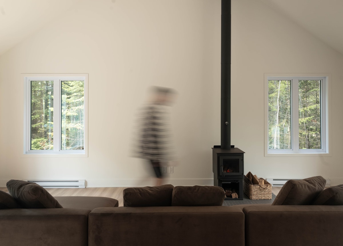 A spacious interior is highlighted by tall ceilings and natural light filtering through large windows. A dark wood stove stands centrally, surrounded by a comfortable sectional sofa. A sense of tranquility is enhanced by the view of the surrounding forest.