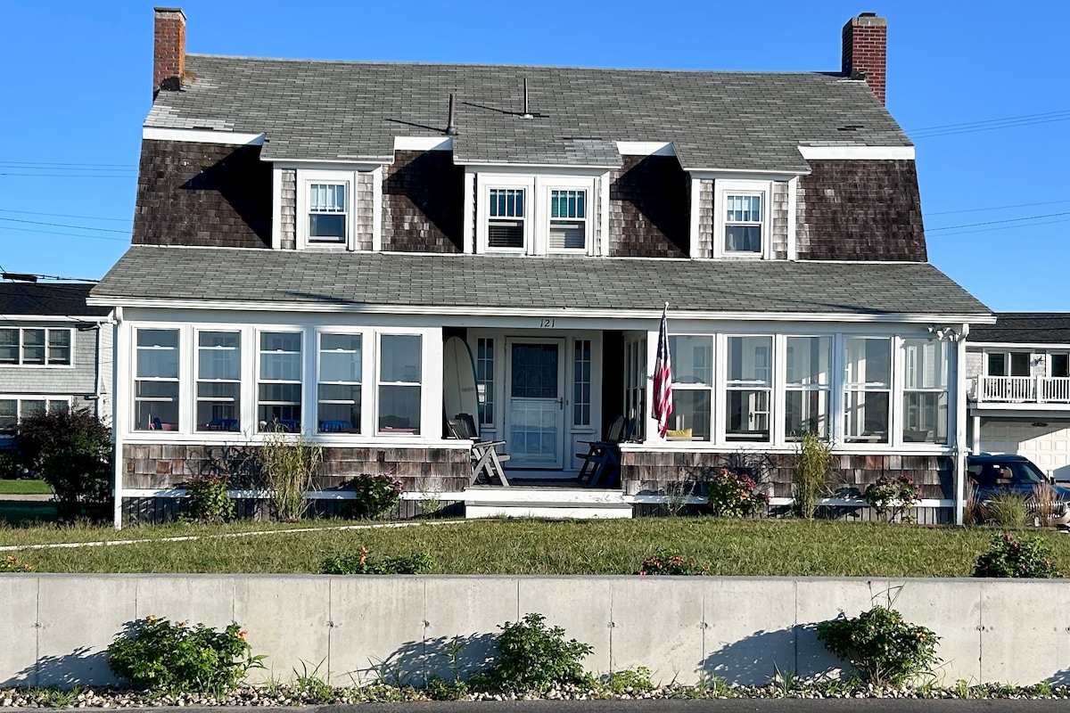 A spacious beachfront home is presented with a distinct gabled roof and shingle siding. The front porch features multiple chairs and is lined with potted plants. An American flag is displayed prominently. Large glass windows allow for ample natural light, offering views of the surrounding landscape.