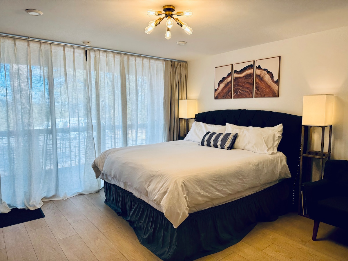 A cozy sleeping area features a spacious bed with crisp white linens and decorative pillows. A striking chandelier hangs above, while large windows with sheer curtains allow natural light to fill the room, creating a bright and airy atmosphere.
