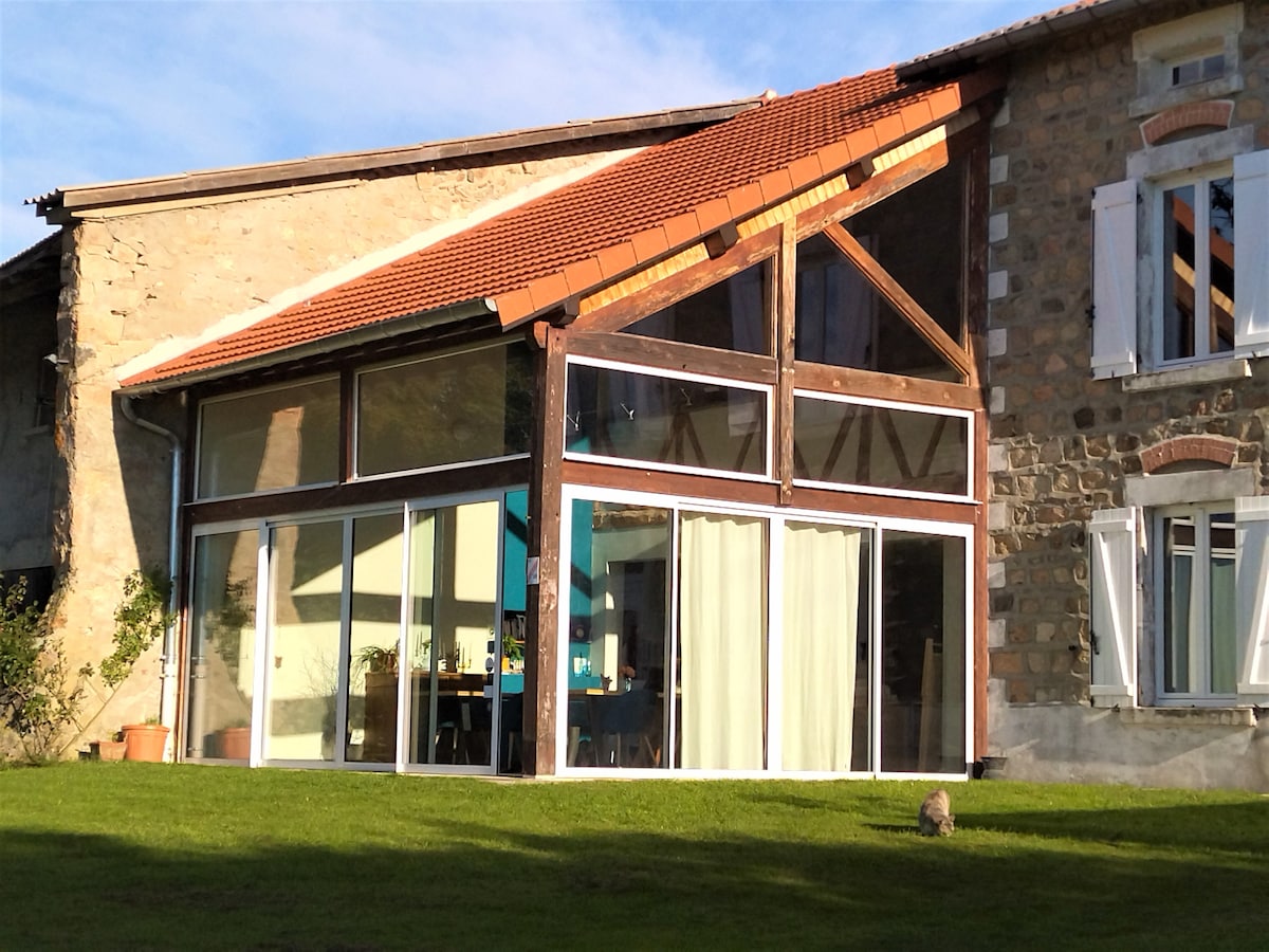 A spacious house is presented with a modern glass facade, featuring large windows that allow natural light to brighten the interior. The structure combines rustic stone walls with contemporary design elements, surrounded by a grassy area and a clear blue sky in the background.