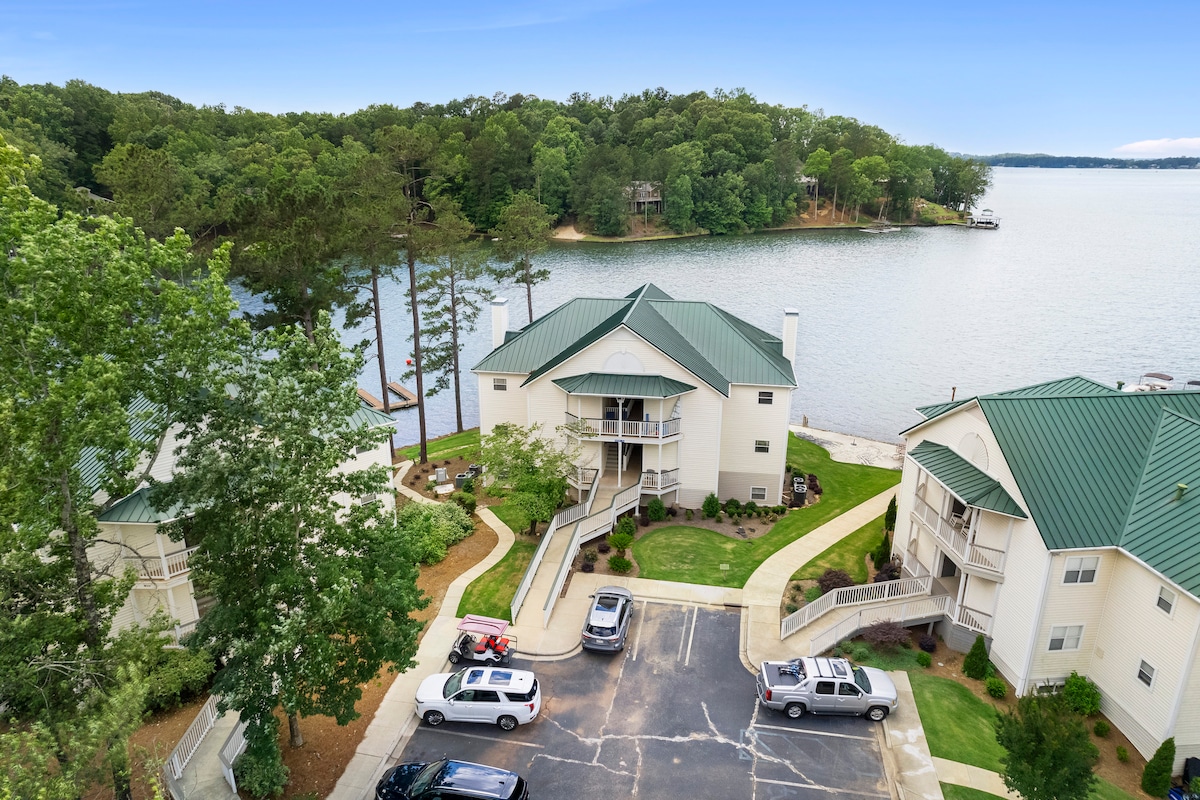 Aerial view of a waterfront condo complex nestled among trees, with a serene lake visible in the background. The image showcases a well-maintained parking area in the foreground, lined with several vehicles, while the landscaping reflects a natural environment.
