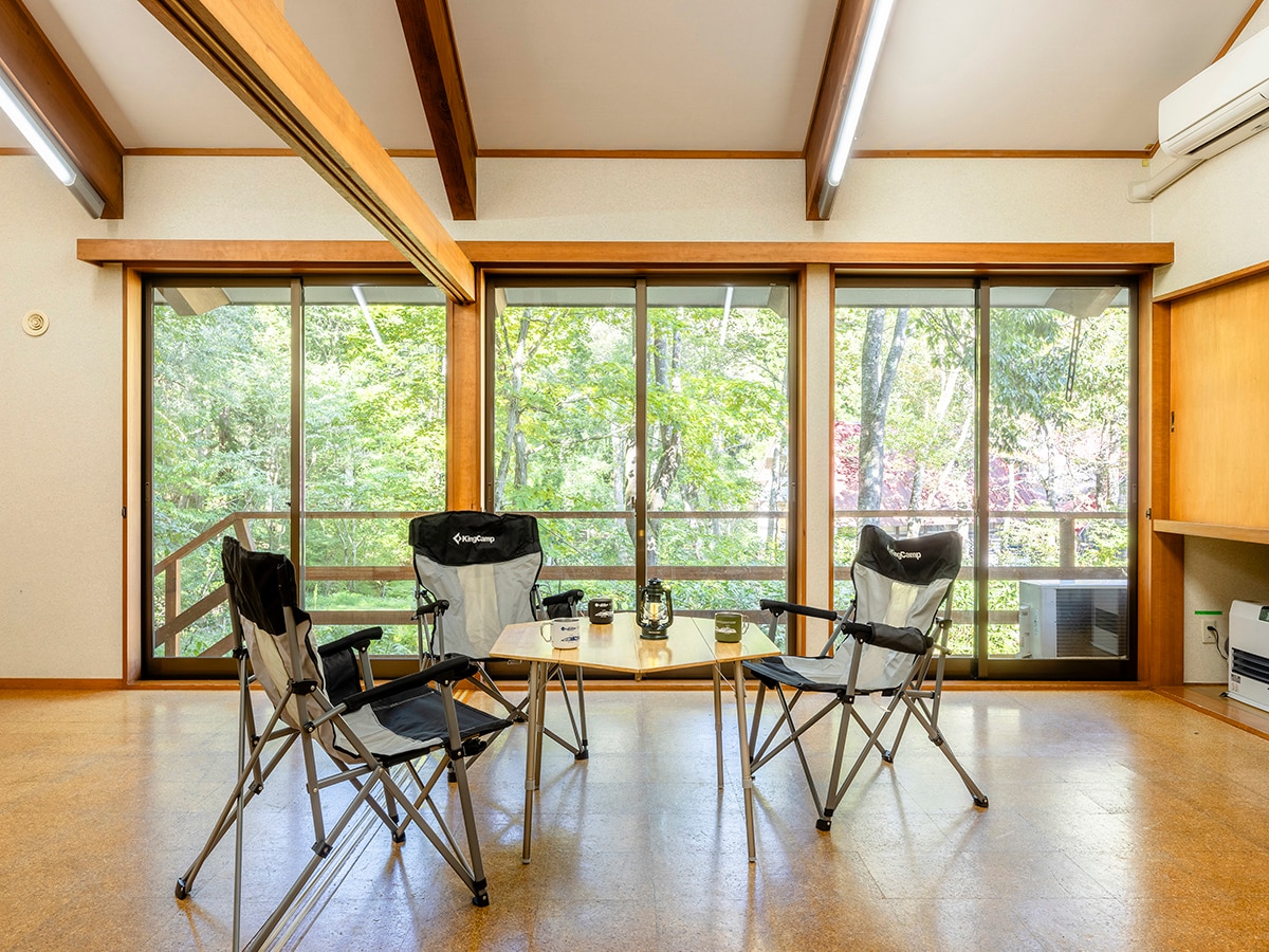 An open room features two folding chairs placed around a small table. Large windows provide a view of the surrounding forest, allowing natural light to brighten the space. Light-colored walls and wooden beams contribute to a spacious and airy atmosphere.