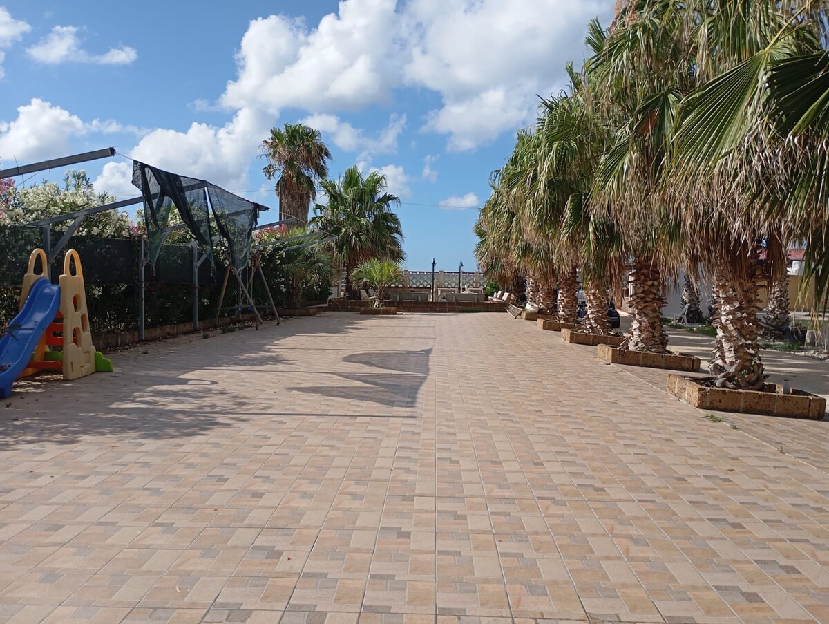 A spacious outdoor area is paved with square tiles, lined with palm trees. A playground structure for children is visible on one side. The scene is brightened by scattered clouds in a blue sky, enhancing the welcoming atmosphere of this communal space.