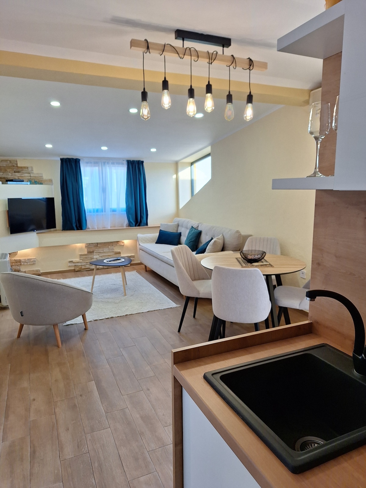 A light-filled living area features a comfortable sofa, accent chairs, and a coffee table. Large windows are adorned with curtains, allowing natural light to illuminate the space. The kitchen area includes a modern sink and cabinetry, creating a cohesive open layout.