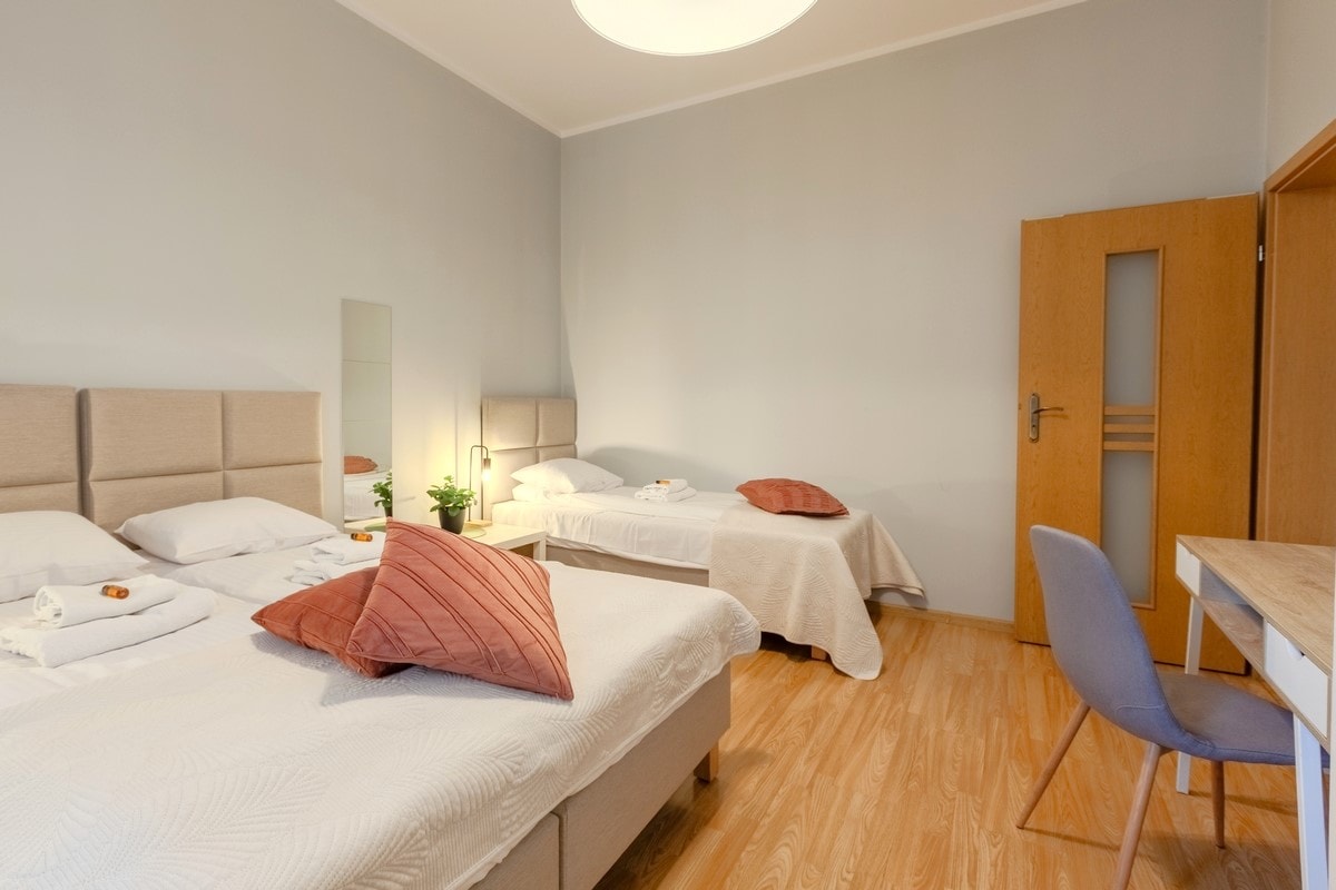 A well-lit bedroom features two double beds, arranged with fresh linen and pillows. A bedside plant adds a touch of greenery. A wooden desk and chair are positioned against the wall, providing a workspace option. Natural light enters through a large window, enhancing the inviting ambiance.