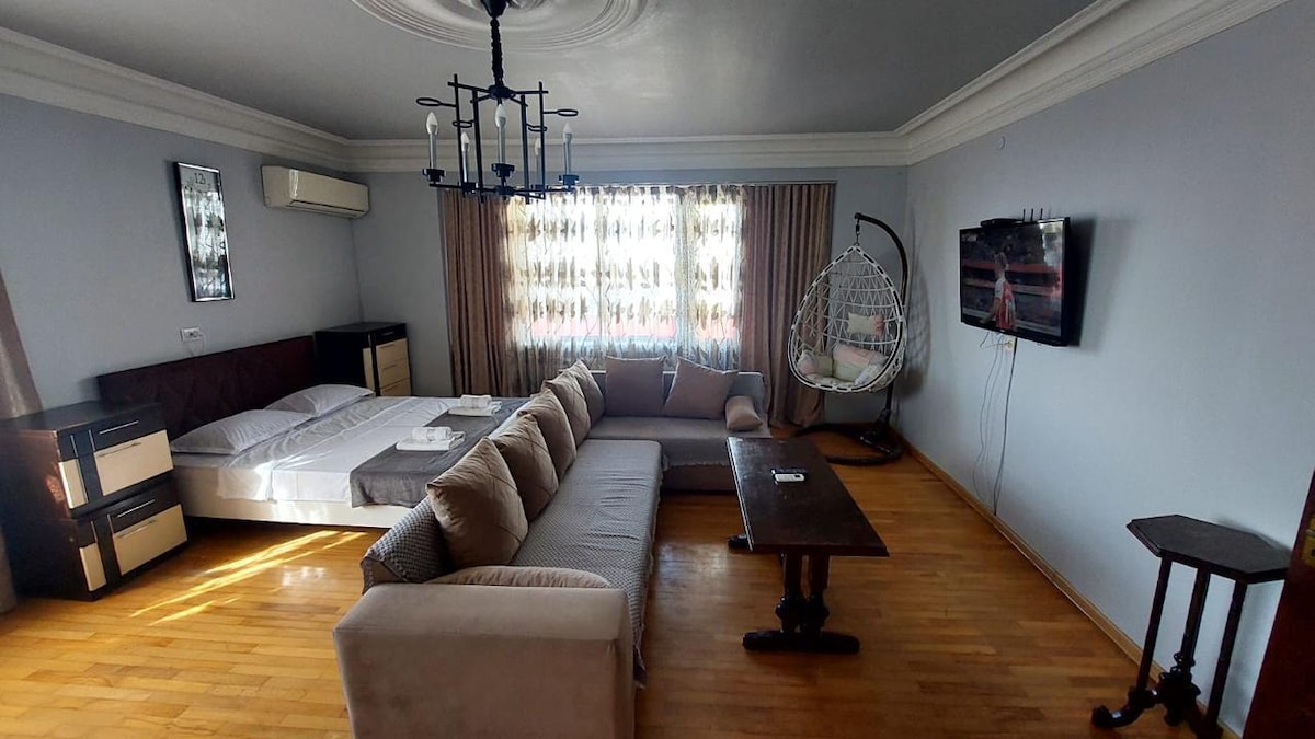 A spacious living area features a modern design with a comfortable sofa and coffee table. A large window with sheer curtains allows natural light to fill the room. A cozy seating nook includes a hanging chair, and a flat-screen TV is mounted on the wall.