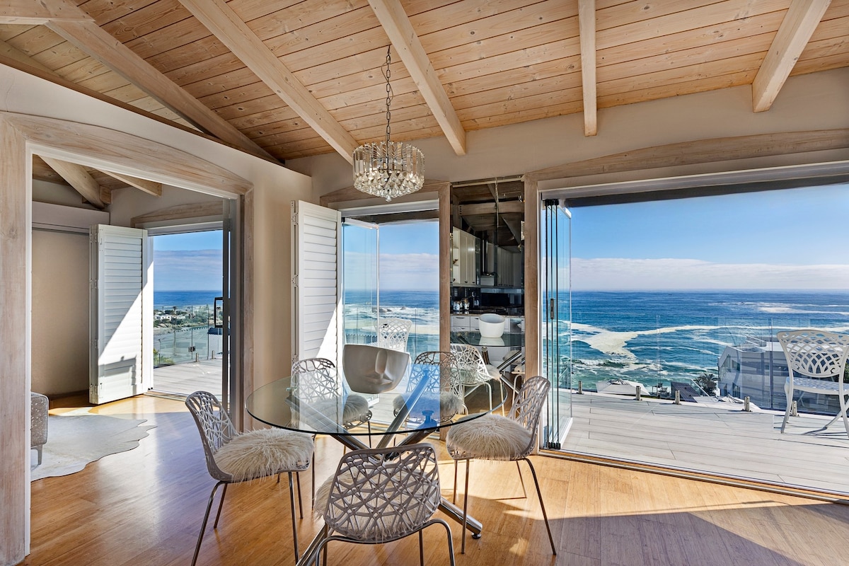 An open dining area features a glass table surrounded by stylish chairs. Expansive sliding doors lead to a balcony offering sweeping ocean views. Natural light fills the space through the large windows framed by light wooden beams.