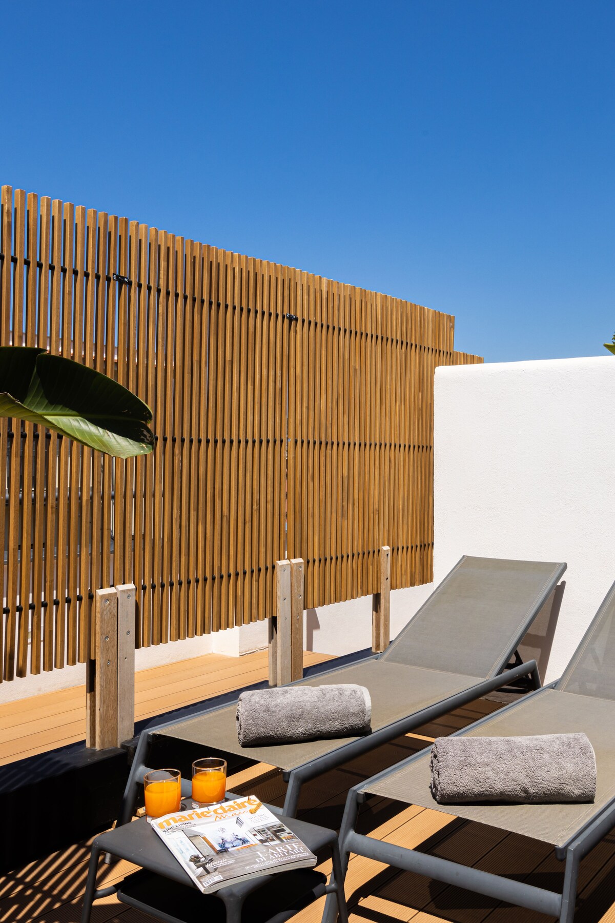 A private solarium is featured, showcasing two loungers with soft towels placed on them. A small table holds two drinks and a newspaper. The area is surrounded by wooden fencing, providing a sense of privacy, while a clear blue sky is seen above.