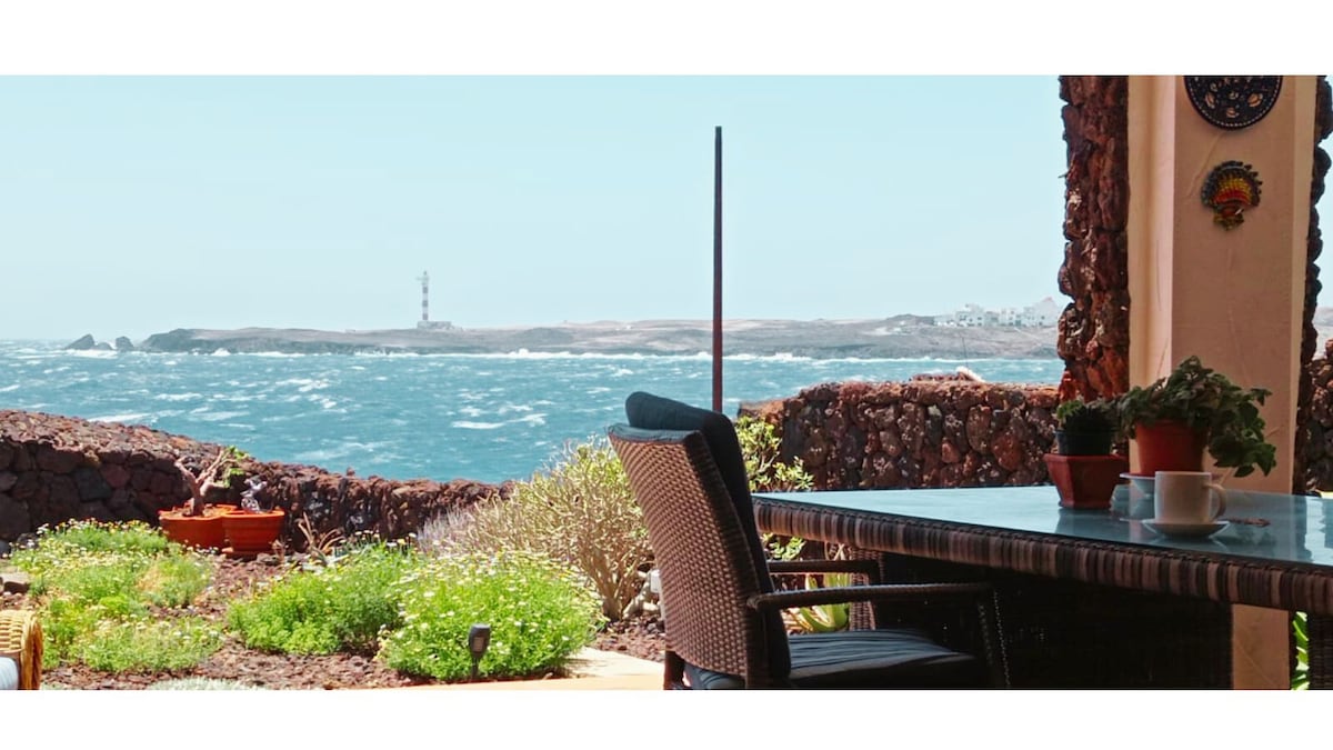 A charming outdoor terrace features a dining table set against a backdrop of the Atlantic Ocean. Coastal vegetation and flowering plants frame the scene, while the distant lighthouse can be seen on the shoreline. The space is designed to blend relaxation with ocean views.