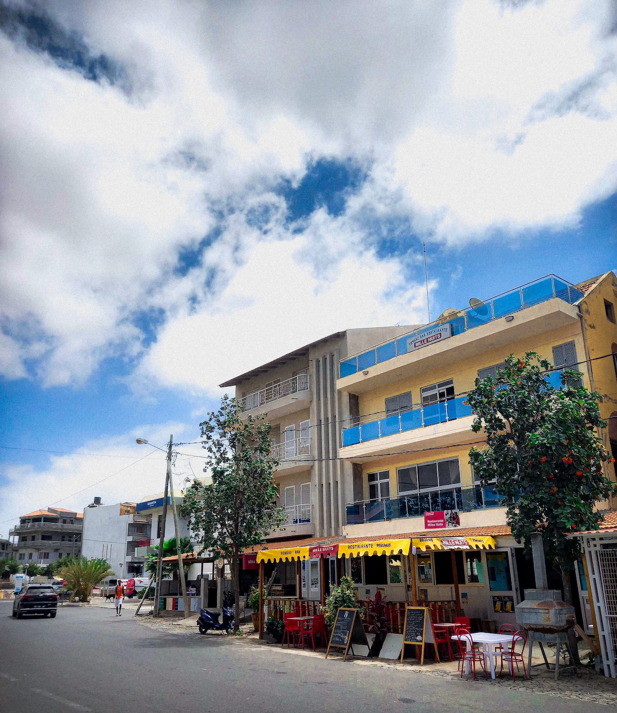 A lively street scene in Tarrafal showcases colorful buildings and outdoor dining areas. Trees are lined along the sidewalk, adding greenery. Bright yellow awnings cover restaurant patios, and vehicles are parked along the street, with a clear blue sky and fluffy clouds overhead.