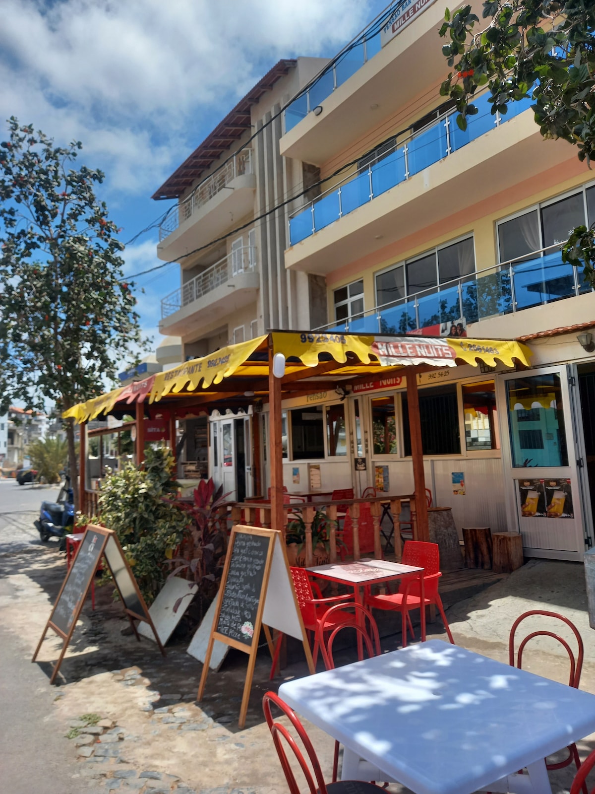 A colorful outdoor seating area is visible under a vibrant yellow awning, with red chairs and tables arranged for diners. The restaurant's menu boards are positioned at the entrance, complemented by greenery. Above, a three-story building shows large windows and a contemporary facade.