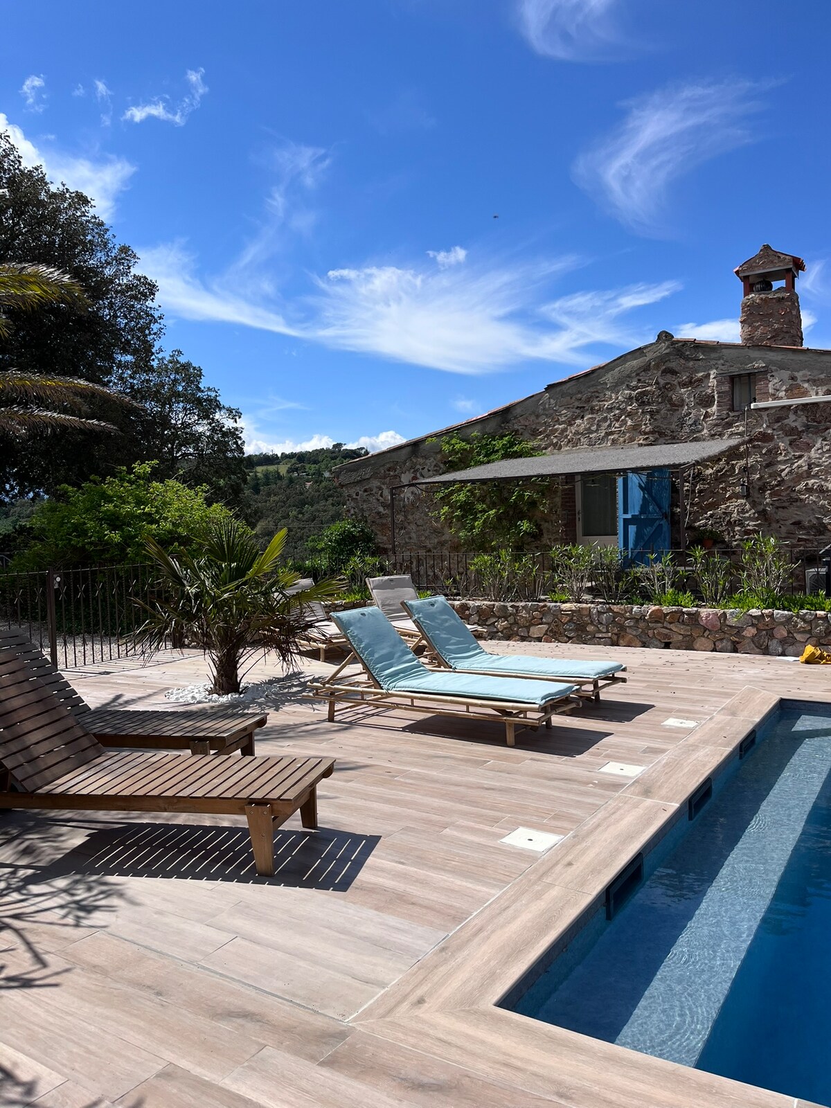Sun loungers are positioned beside a rectangular pool, set against a backdrop of a stone building featuring blue shutters. Lush greenery and flowering plants surround the area, with a clear sky overhead and wispy clouds adding to the serene outdoor setting.