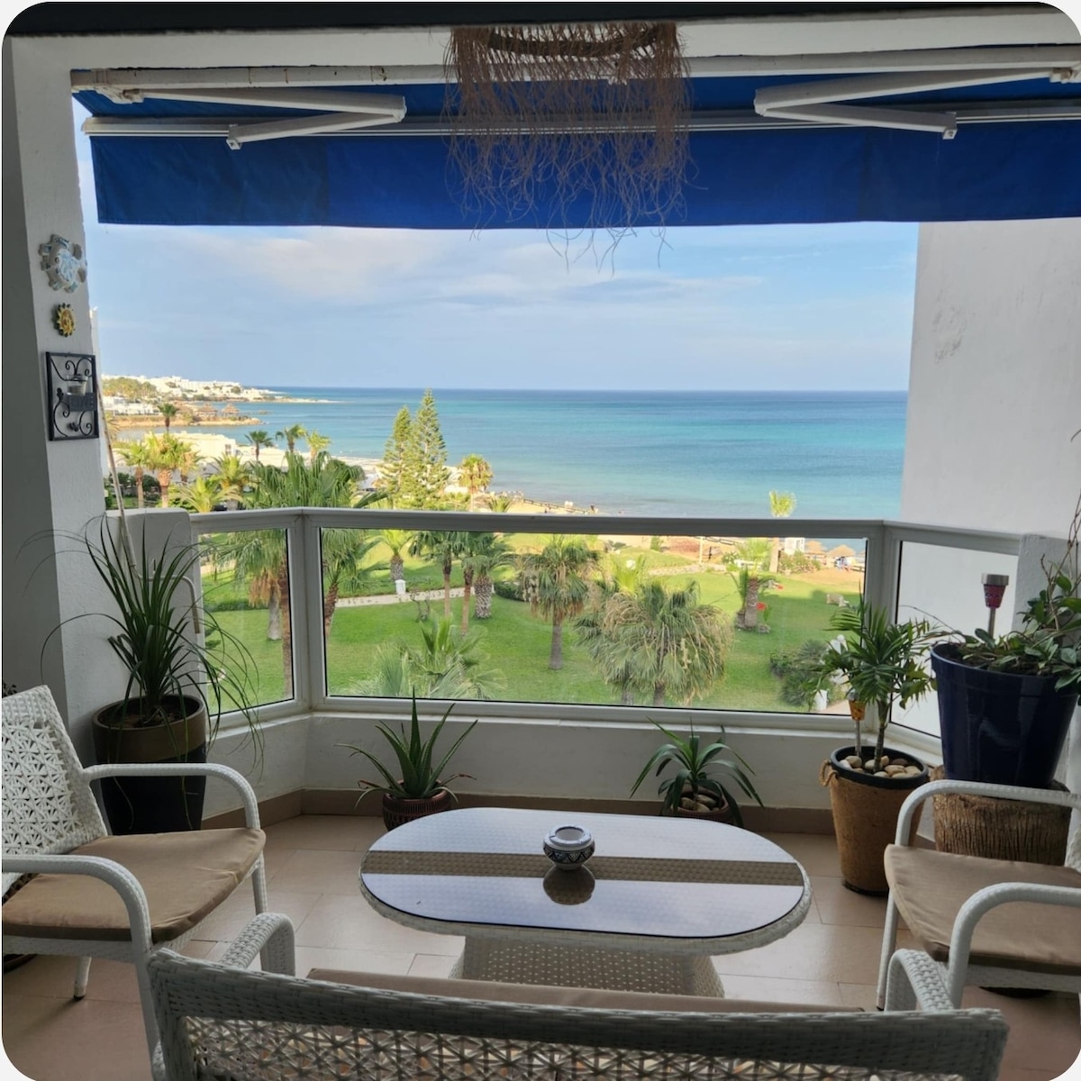 A serene terrace is showcased, featuring a glass railing that provides an unobstructed view of the beach and lush garden below. Comfortable seating is arranged with a round coffee table at the center, surrounded by potted plants that add a touch of greenery.