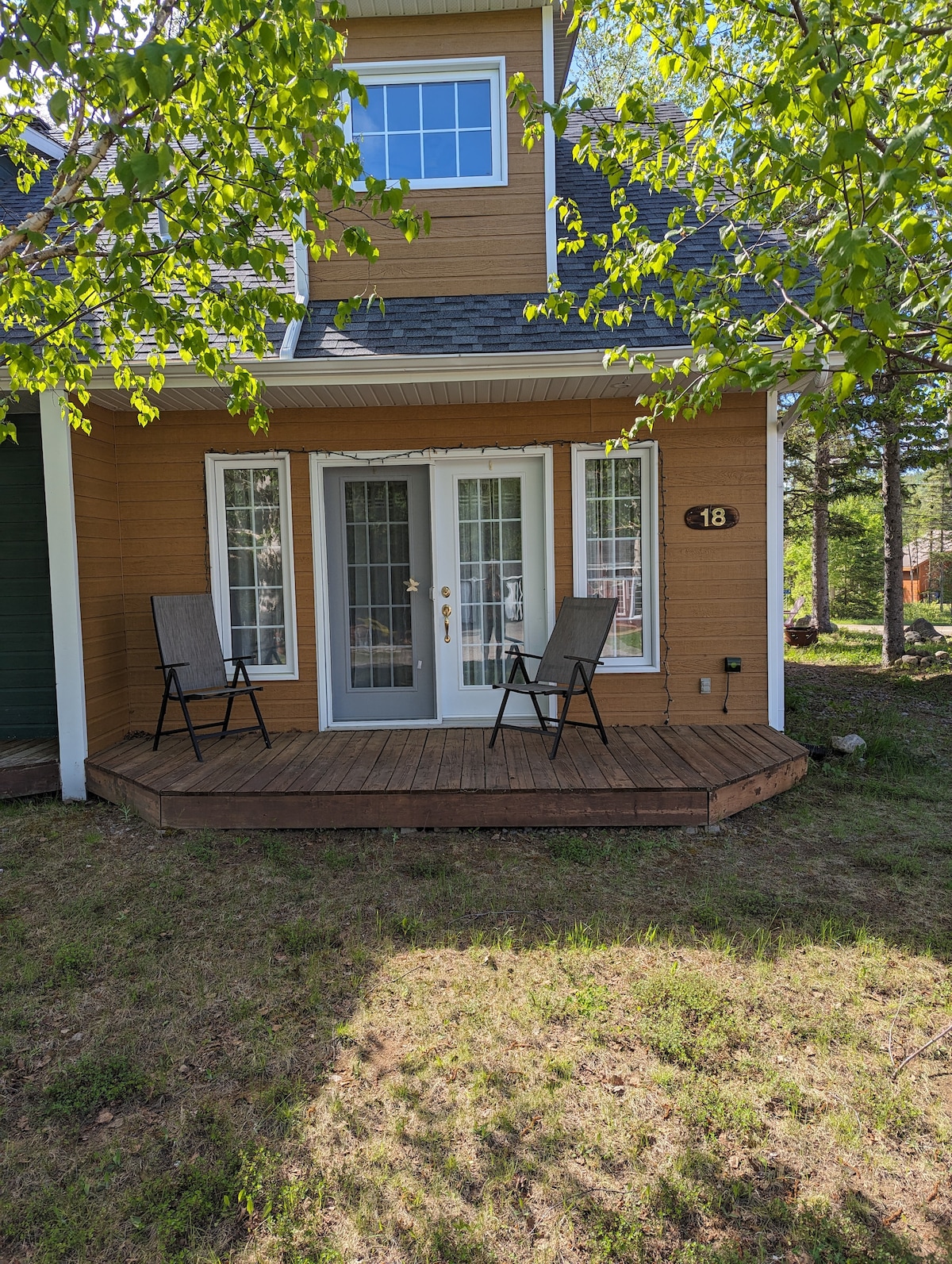 A welcoming exterior features a wooden deck with two chairs positioned invitingly. Light wood siding complements the surroundings, and large glass doors create a connection to the indoors. Soft greenery frames the scene, enhancing the tranquil setting.