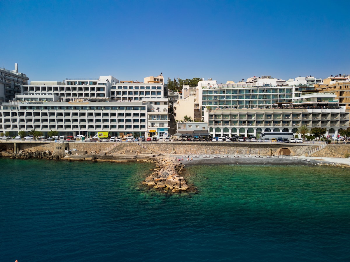 A seafront building is depicted, showcasing a modern architectural design with multiple levels. The structure features balconies facing the water, while the clear turquoise waters lap against a small rocky jetty. A beach with sun loungers is positioned nearby, creating a relaxed coastal atmosphere.