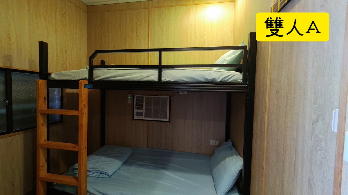 A cozy sleeping area is shown with a bunk bed and a spacious bed below. Light blue bedding is neatly arranged. An air conditioning unit is mounted on the wall, and wooden walls create a warm ambiance.