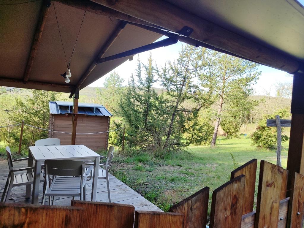 An outdoor terrace is shown with a table and four chairs, surrounded by a natural landscape. In the background, a wooden structure is visible, which serves as an outdoor toilet. Lush greenery and trees create a peaceful setting.