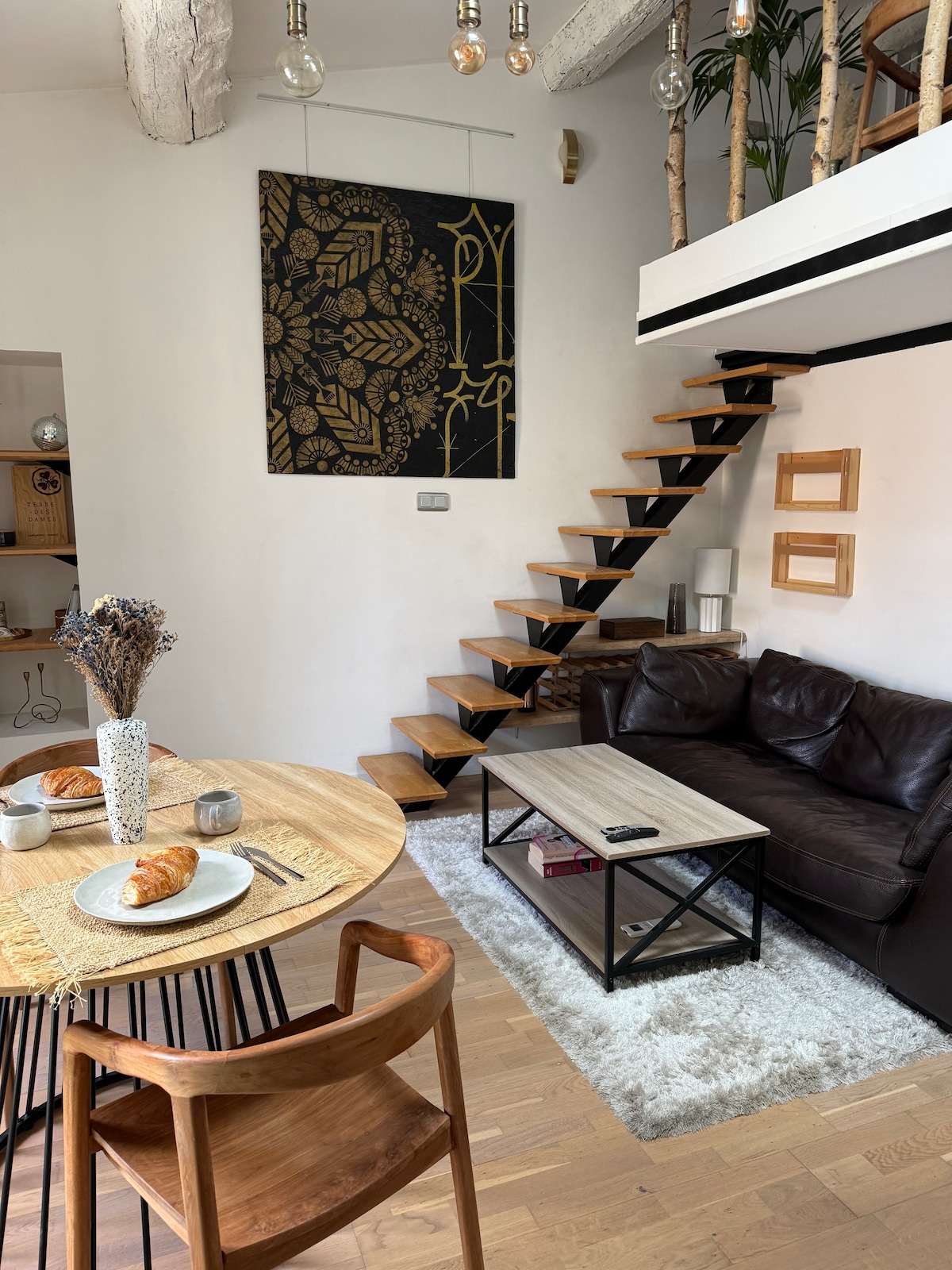 An inviting living space features a wooden dining table set with plates and pastries. A brown leather sofa complements the decor, while a textured rug adds warmth. Decorative elements include a wall art piece and a staircase crafted from wood, leading to an upper level.
