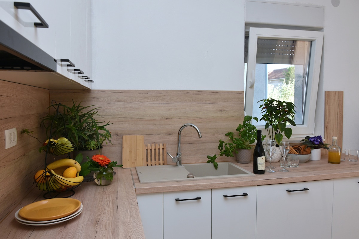 A clean kitchen space features a wooden countertop and white cabinetry. Potted plants are arranged on the counter, alongside a sink and several glassware pieces. A bottle of wine and plates with fresh fruits are positioned neatly, offering a functional yet inviting culinary area.