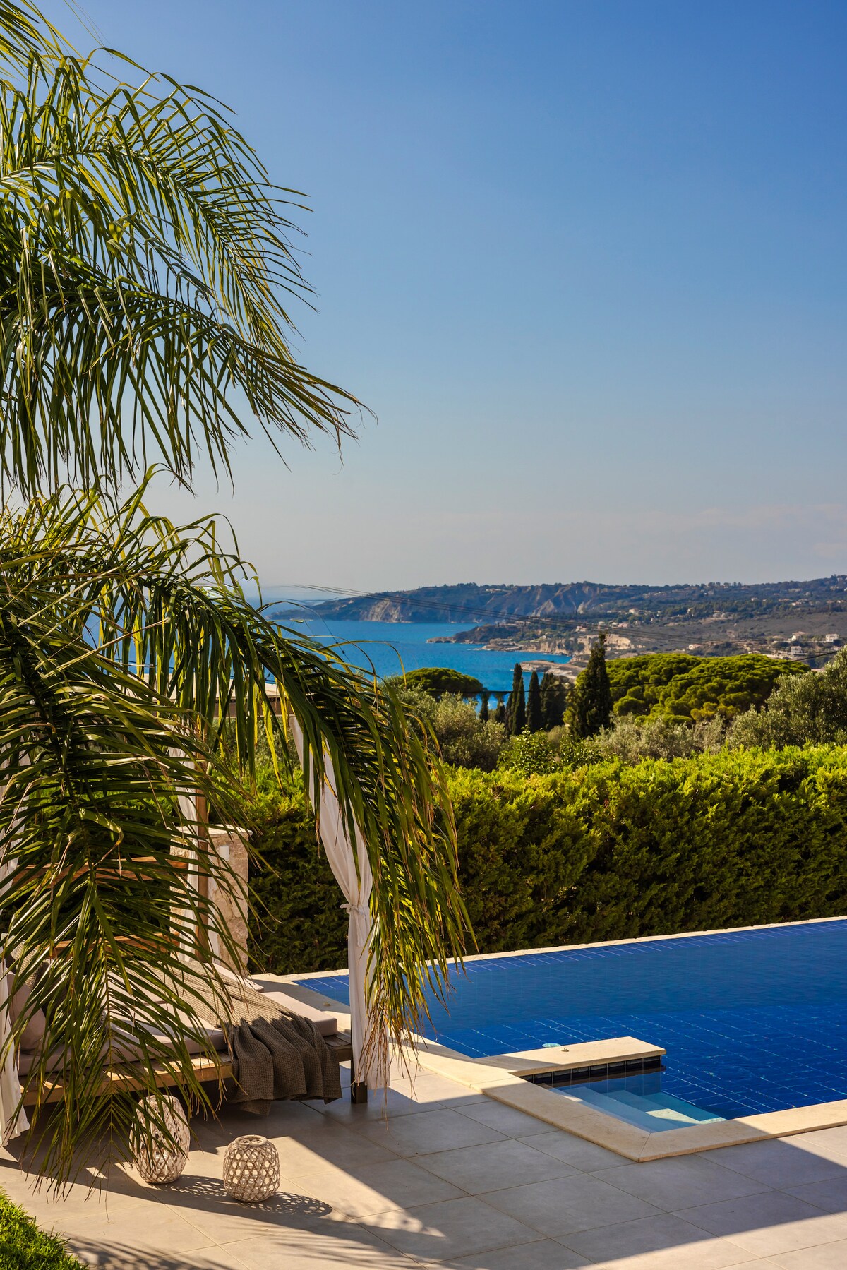 A serene outdoor space features a private pool with serene blue tiles against a backdrop of the Ionian Sea. Lush greenery and tall palm trees frame the area, enhancing the tranquil atmosphere. Clear skies provide ample sunlight, reflecting off the water's surface.