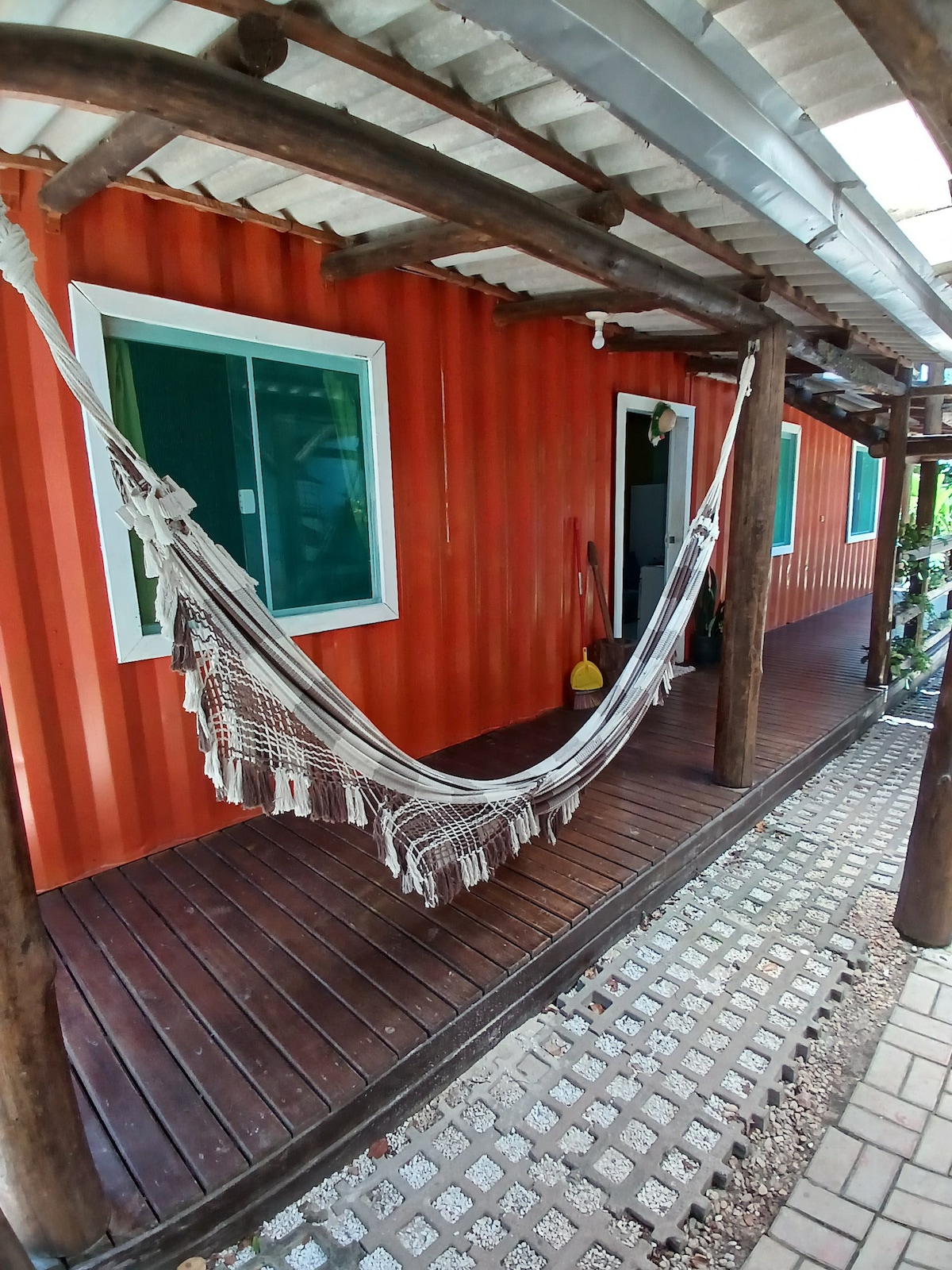 A shaded outdoor area features a wooden deck with a decorative hammock hanging between two rustic posts. The entrance of a bright orange container home is visible, complemented by a large window allowing natural light to fill the space.