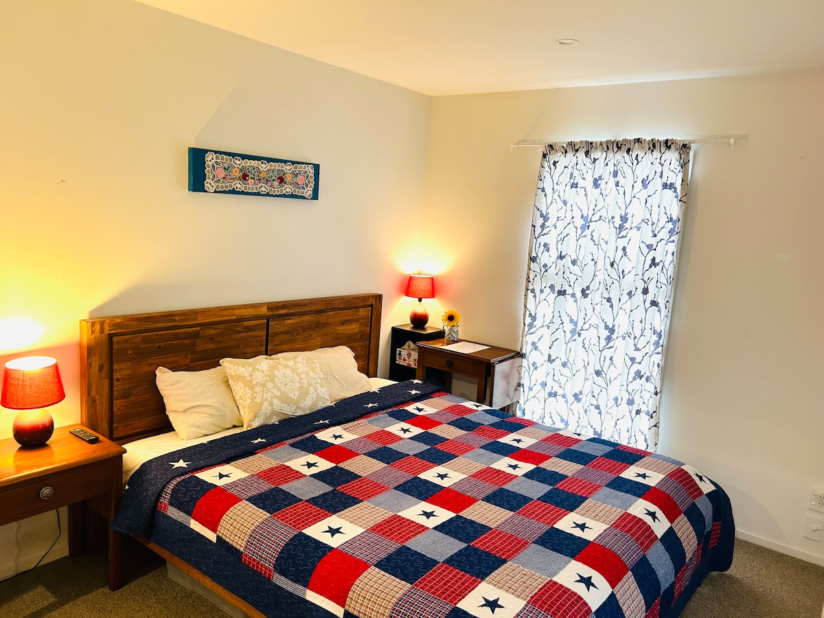 A bedroom features a spacious bed covered with a blue and red plaid quilt. Two bedside tables with lamps stand beside the bed. A window with light-filtering curtains allows soft light to enter, complementing the neutral-toned walls.