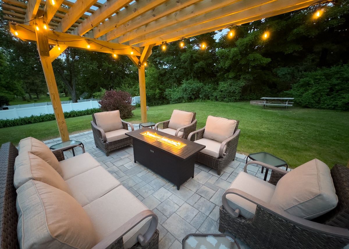 An outdoor seating area is arranged under a wooden pergola adorned with string lights. Comfortable chairs surround a central fire table, which is equipped for warmth and ambiance. Lush greenery and a picnic table are visible in the background, contributing to the serene outdoor setting.