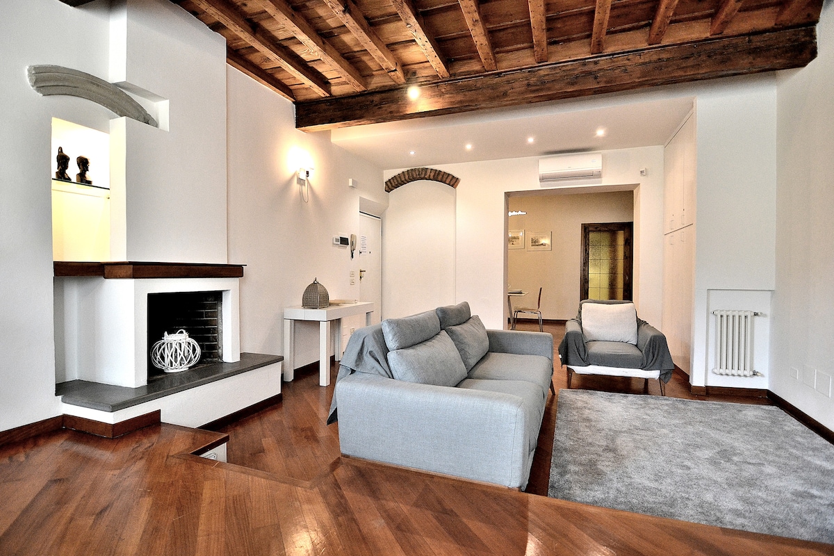 A spacious living area is showcased, featuring a light grey sofa and an armchair, complemented by a polished wooden floor. A simple fireplace is positioned against a white wall, and warm recessed lighting enhances the overall ambiance.