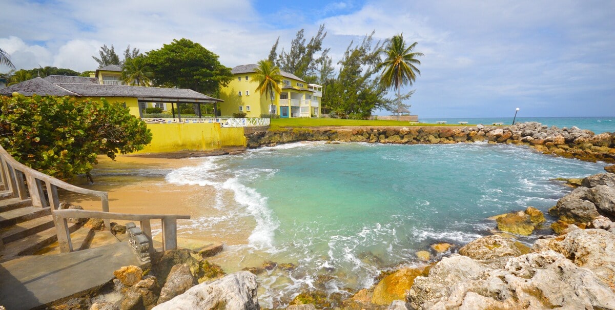 The shoreline features gentle waves lapping against smooth rocks, with tree-lined greenery providing a serene backdrop. A yellow building is visible in the distance, harmonizing with the landscape. The calm waters reflect the sky, enhancing the picturesque coastal setting.