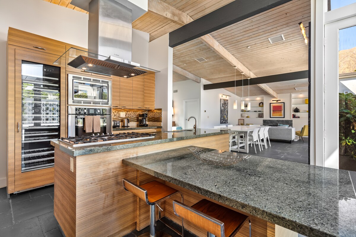 The gourmet kitchen features sleek granite countertops and wood cabinetry. There is a stainless steel oven and wine fridge, as well as bar seating for four. An open layout connects the kitchen to the living area, showcasing a spacious, inviting design.