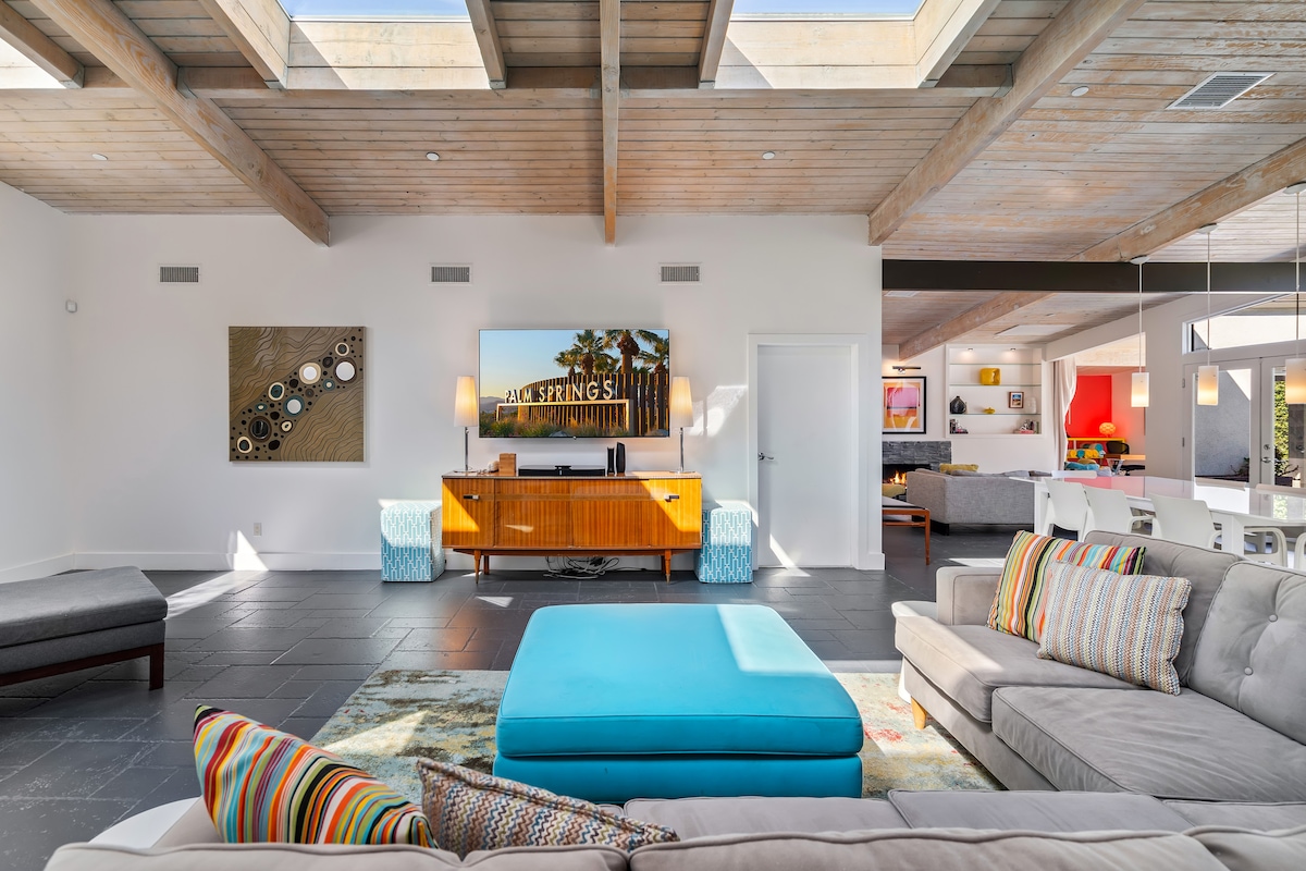 An open living space features a spacious gray sectional sofa, a colorful ottoman, and a large wall-mounted TV. Light pours in from skylights, highlighting midcentury design elements like natural beams and a sleek wooden media console. Artwork adorns the walls, adding a touch of character.
