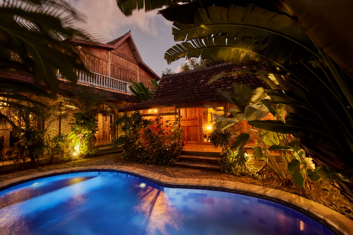 A tranquil pool area is surrounded by lush tropical plants, offering a peaceful retreat. Soft lighting enhances the natural beauty, while traditional wooden buildings frame the space, creating an inviting atmosphere for relaxation.