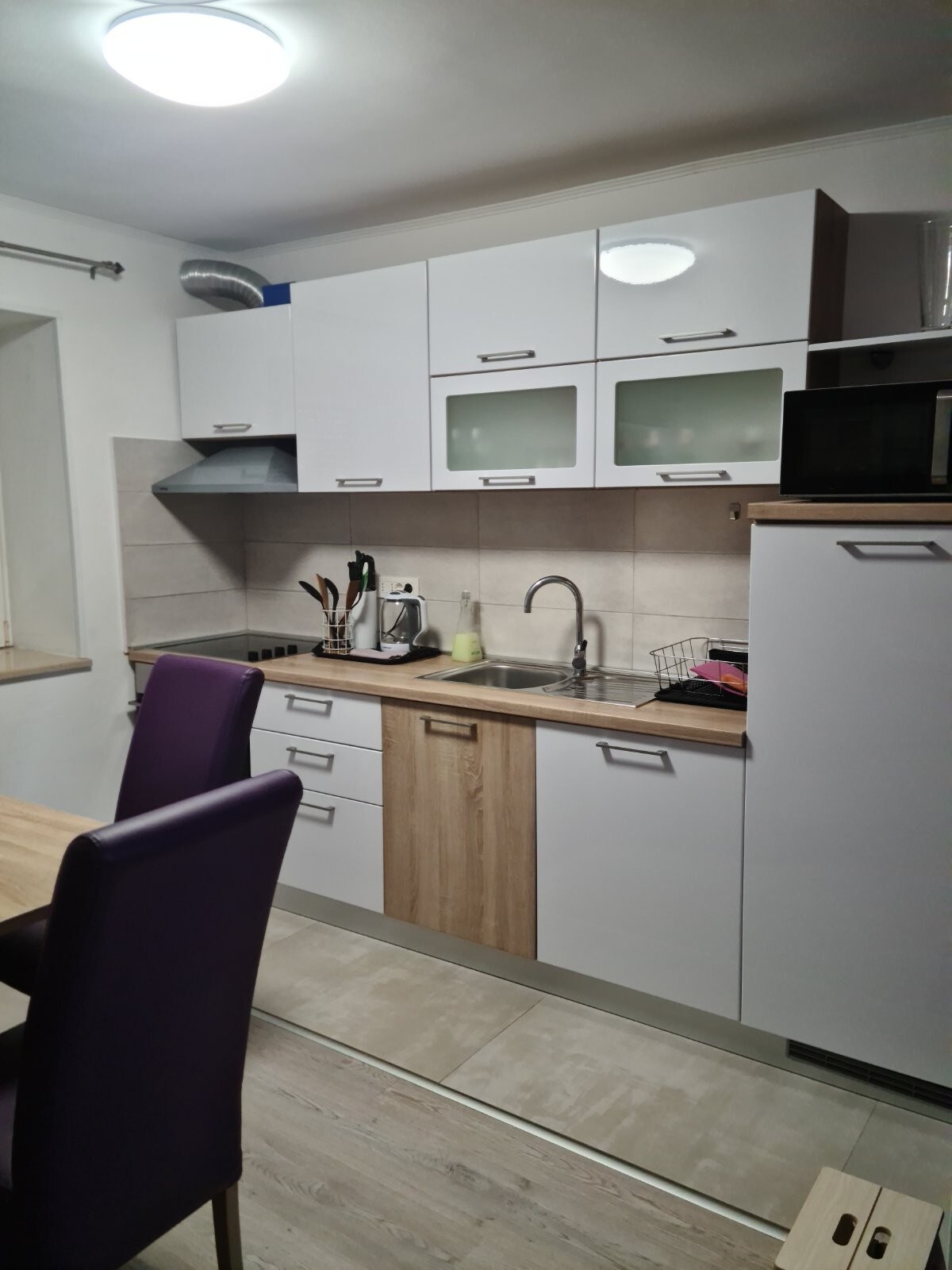 A modern kitchen showcases sleek cabinetry with a mix of white and wood finishes. Kitchen appliances, including a kettle and a microwave, are visible on the countertop. A dining table with purple chairs complements the space, enhancing its functionality.