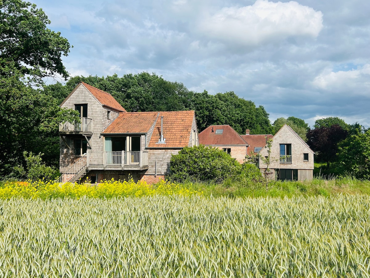 Two charming lodges are nestled among lush greenery, surrounded by a vibrant field of golden crops. The structures feature natural wood accents and red-tiled roofs. A wooden balcony provides an outdoor space, enhancing the connection to the scenic landscape.