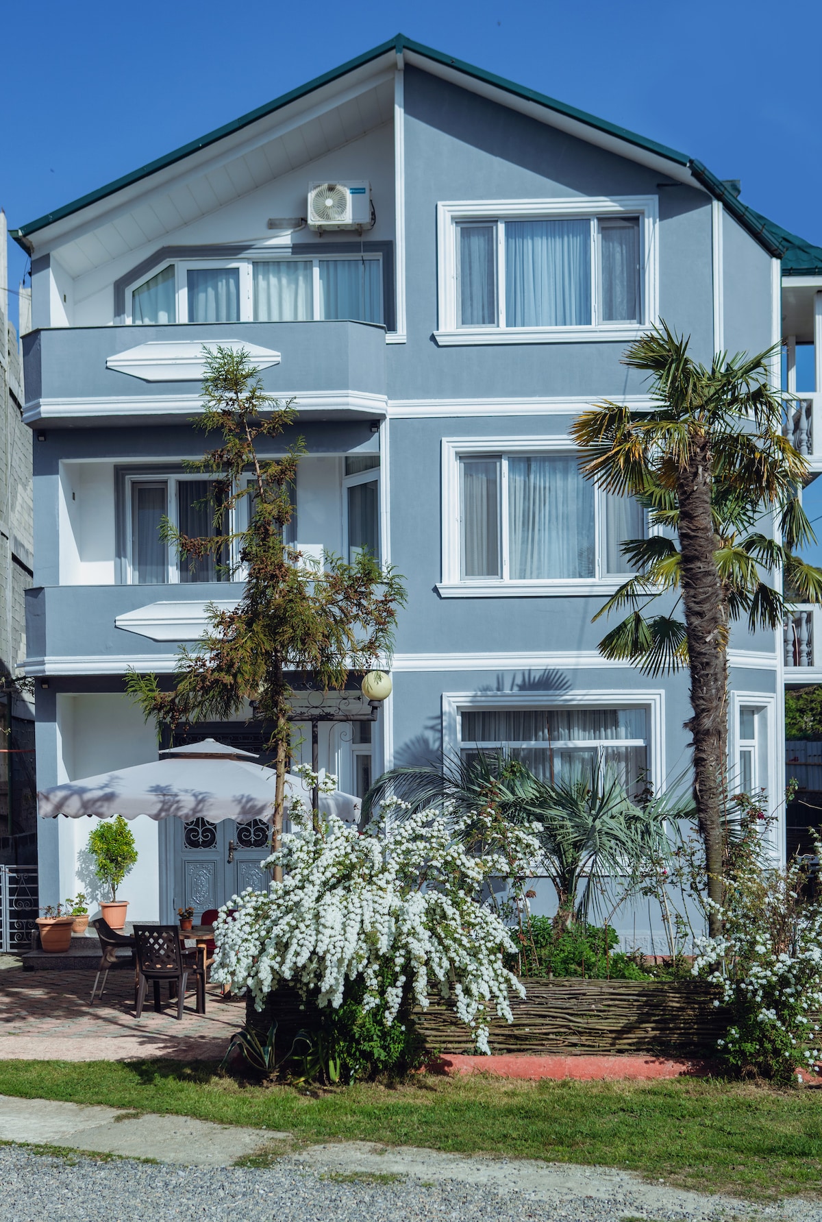 The three-story house features a light blue exterior with multiple large windows. Lush greenery and flowering plants surround the entrance, where a seating area under an umbrella provides a relaxing spot. The clear sky and vibrant foliage enhance the welcoming atmosphere.